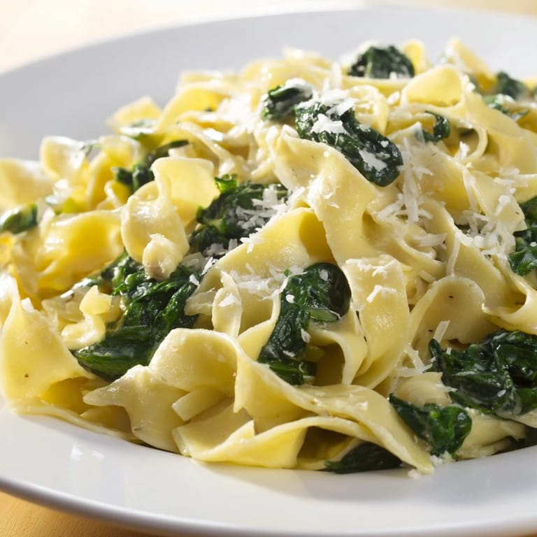 Platter of Creamy Spinach Garlic Noodles garnished with grated Parmesan, next to a glass of crisp white wine on a rustic table.